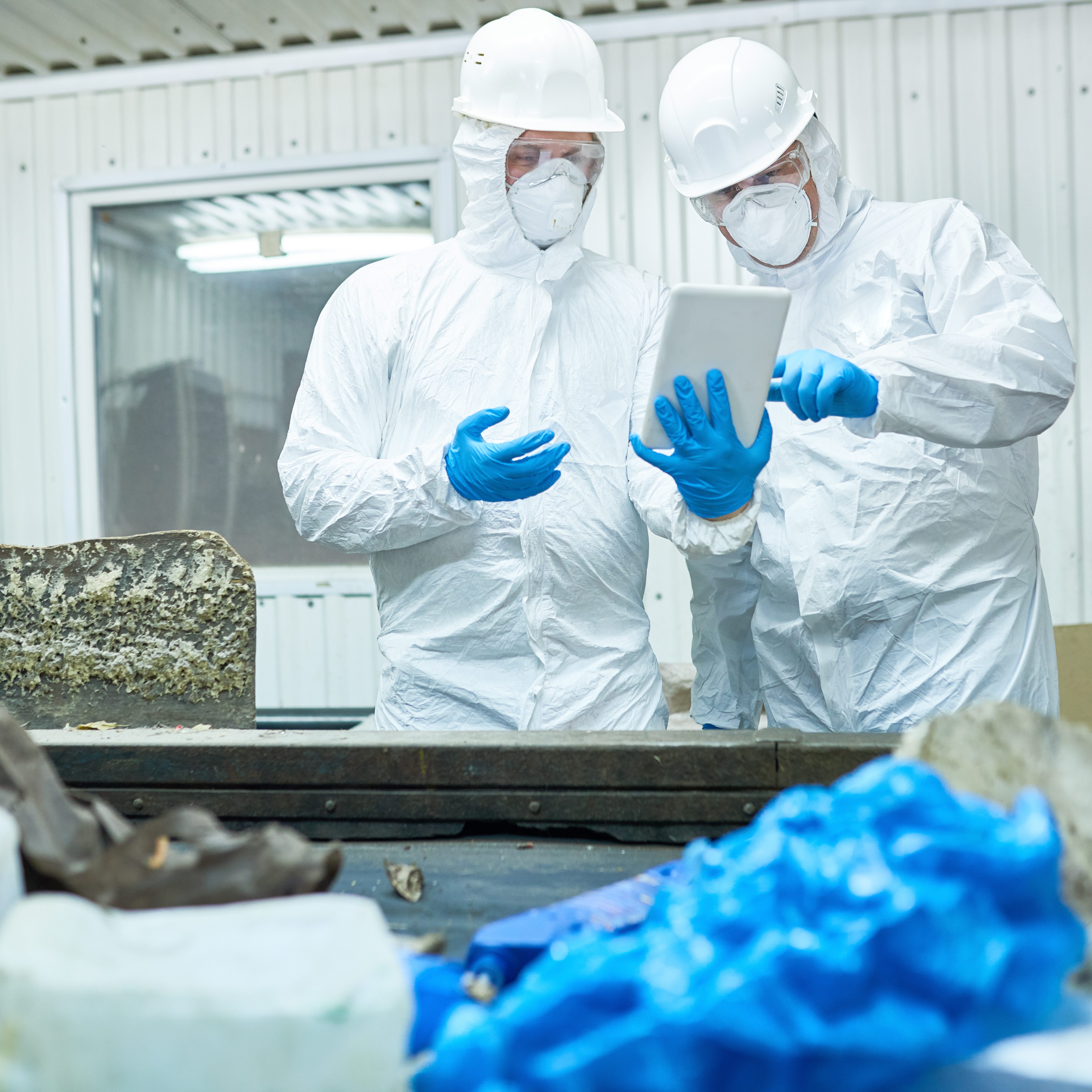 Portrait of two workers  wearing biohazard suits working at waste processing plant using digital tablet and sorting trash standing by conveyor belt, copy space